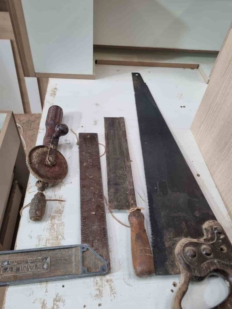 Sobre Mim 3 Quatro ferramentas manuais antigas de marcenaria de mais de 100 anos dispostas sobre uma bancada branca de oficina. À direita, um serrote grande de tração com lâmina de aço escura e cabo de madeira trabalhado. No centro, um serrote de costa menor e um esquadro metálico Stanley em formato de L com pátina de oxidação. Na esquerda, uma furadeira manual de manivela com engrenagens visíveis e ferrugem natural.
